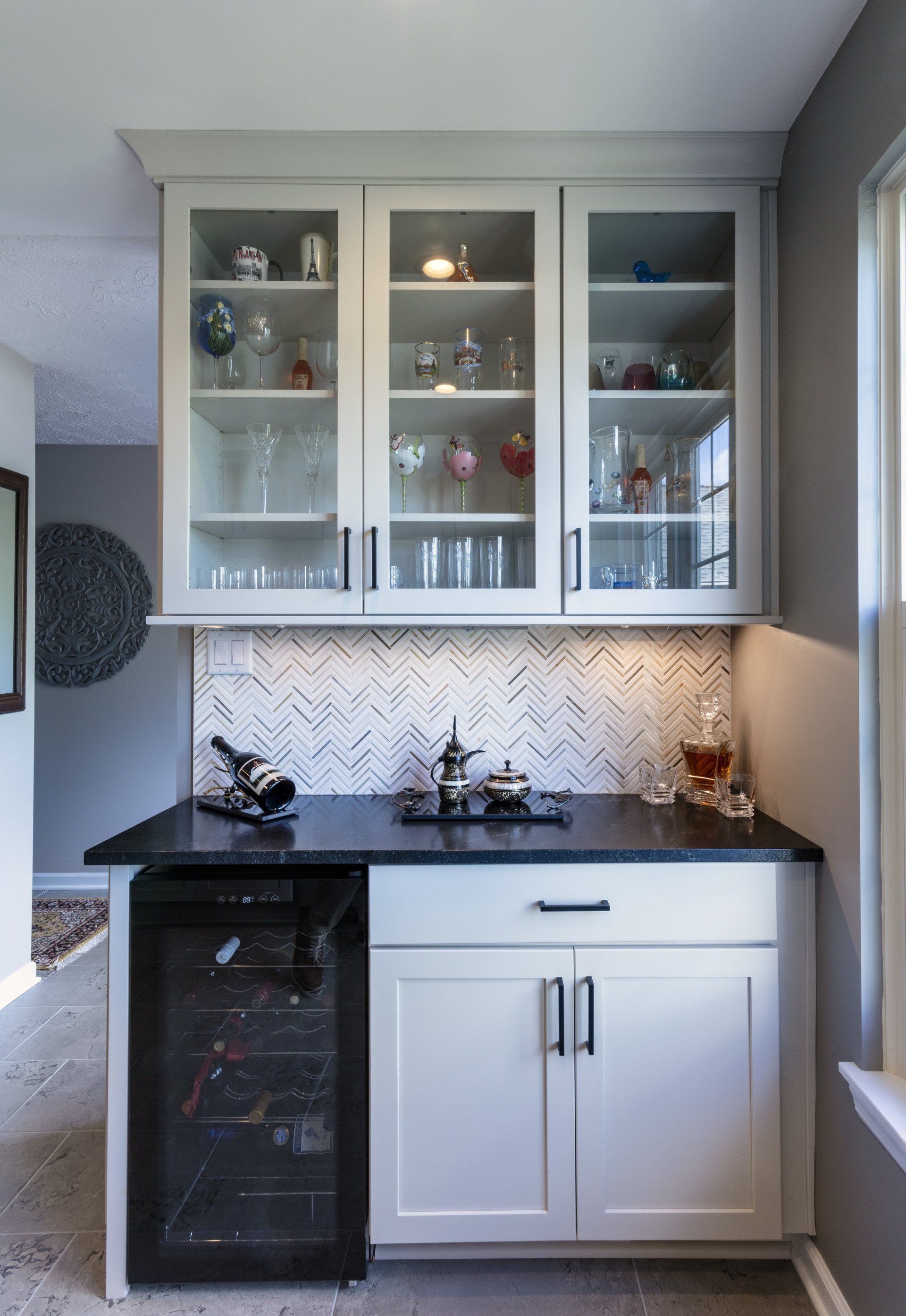 Compact kitchen nook with white cabinets, glass-front hutch, black countertop, and herringbone tile backsplash.