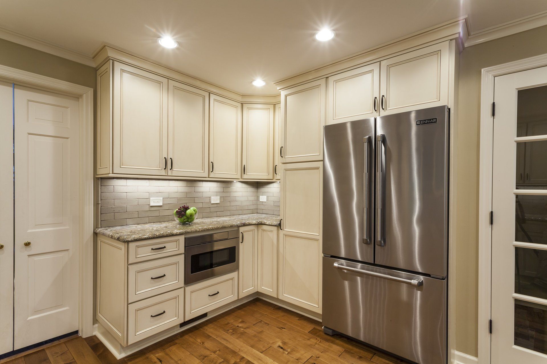 Bright kitchen with white cabinets, stainless steel refrigerator, and built-in oven under recessed lights