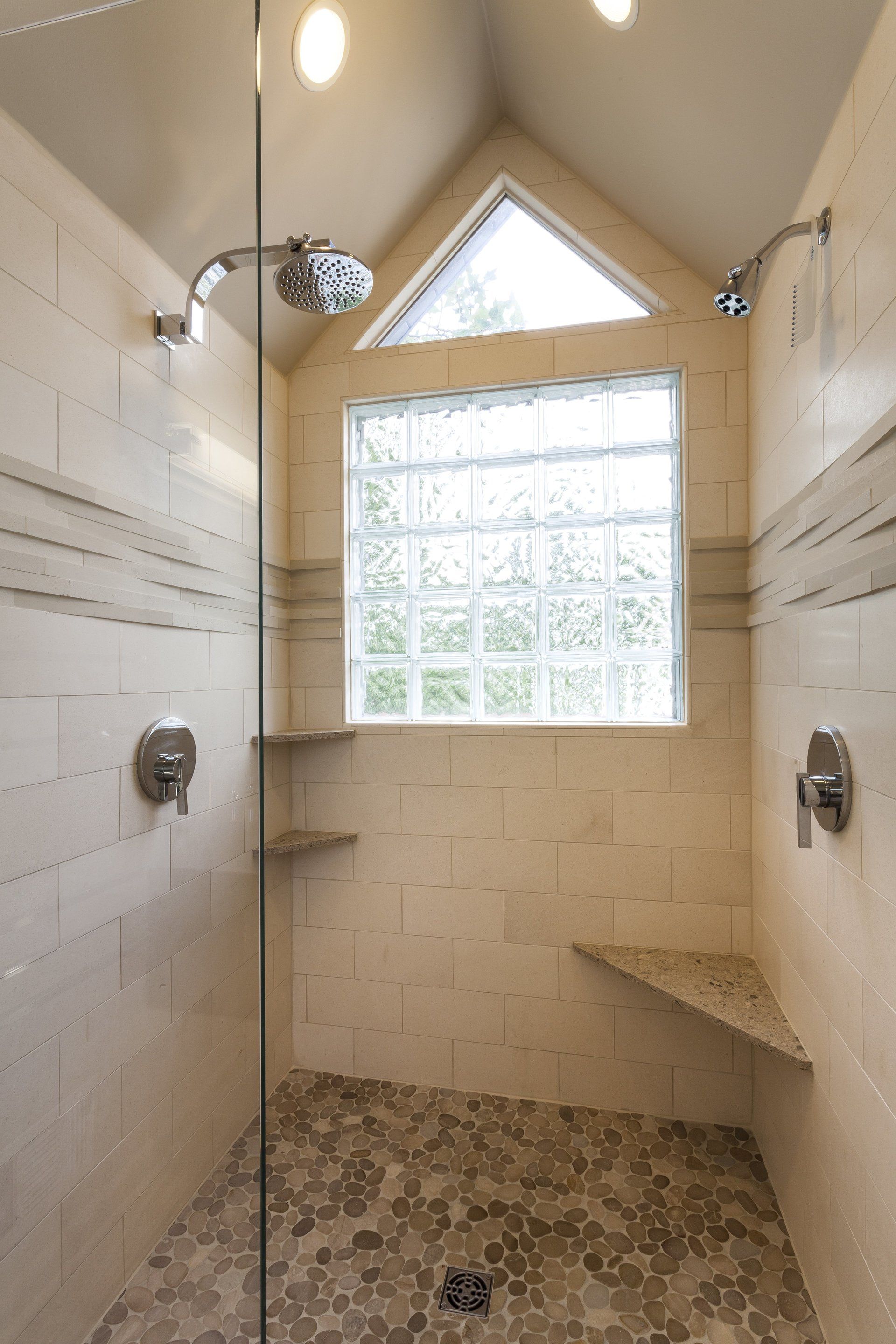 Small tiled shower with glass-block window, triangular skylight, and pebble stone floor.
