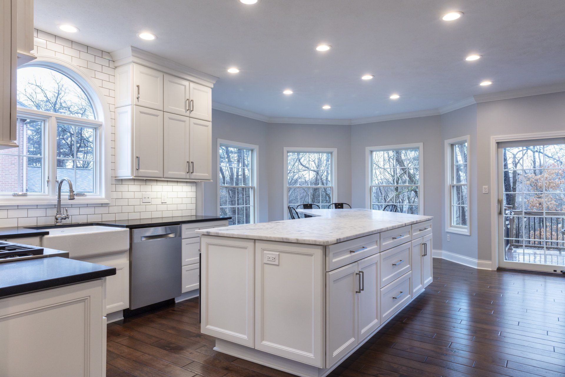 Bright white kitchen with island, dark wood floors, stainless dishwasher, and large windows