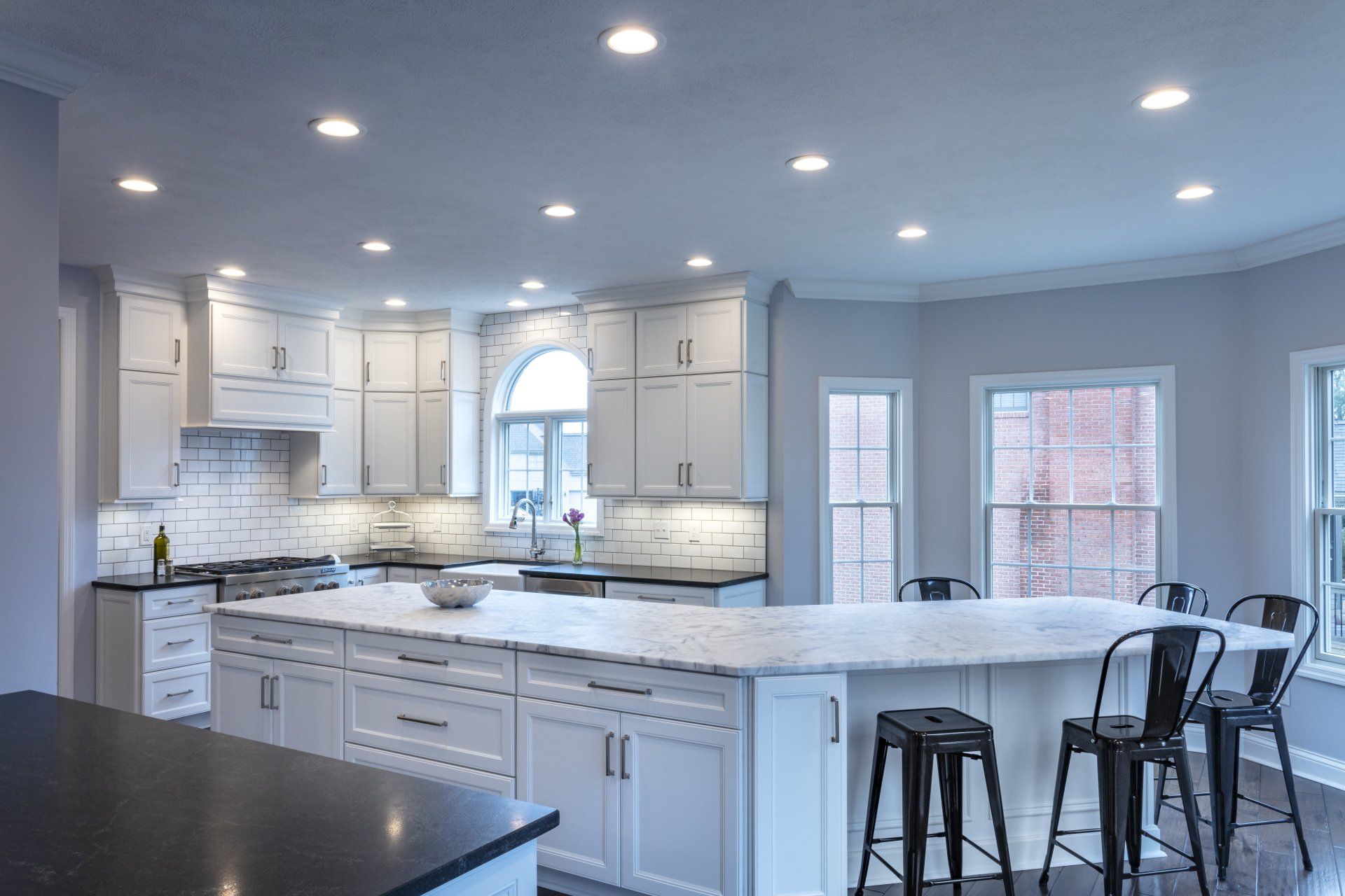 Bright modern kitchen with white cabinets, marble island, bar stools, and recessed ceiling lights.