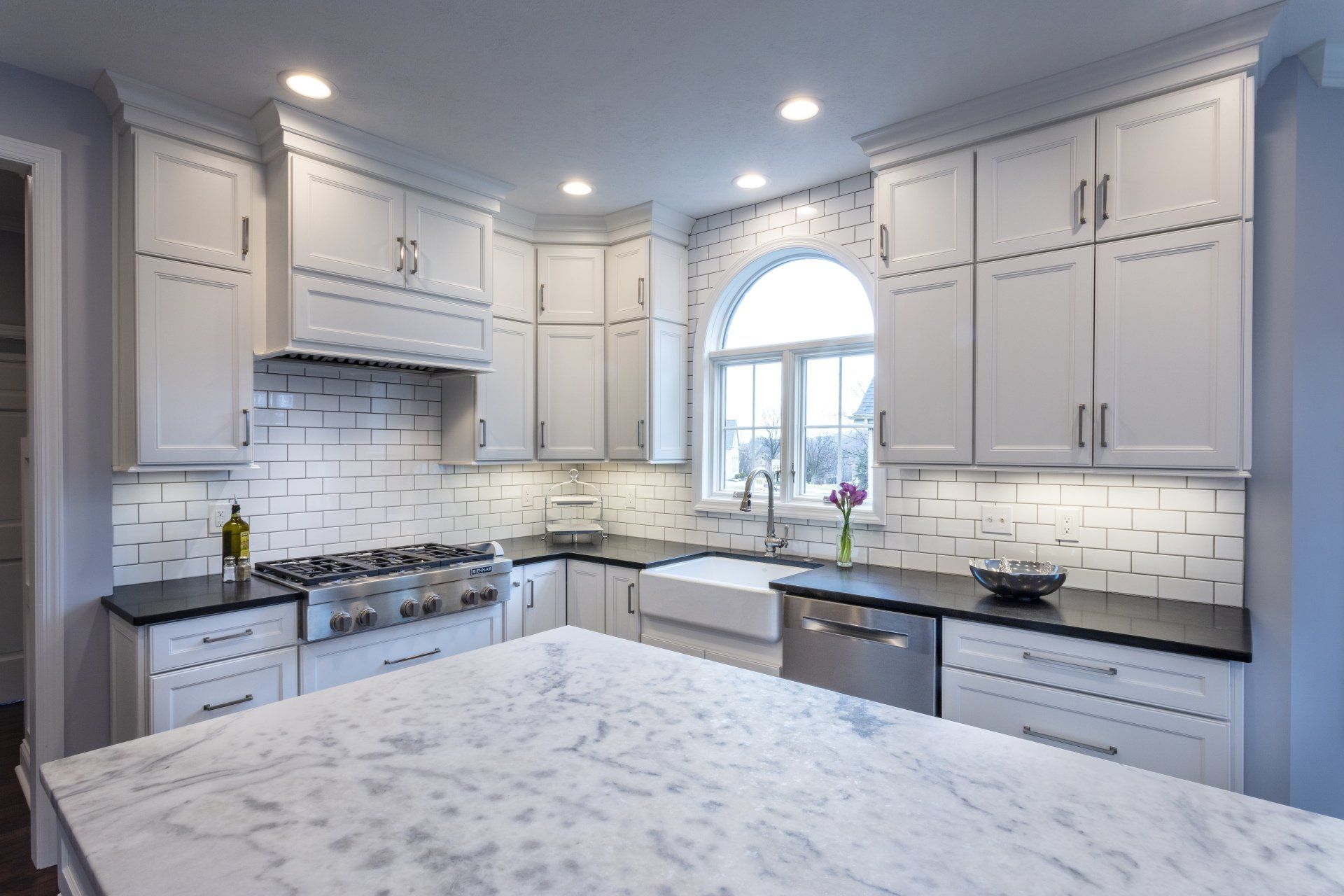 Bright modern kitchen with white cabinets, marble island, and arched window over the sink.