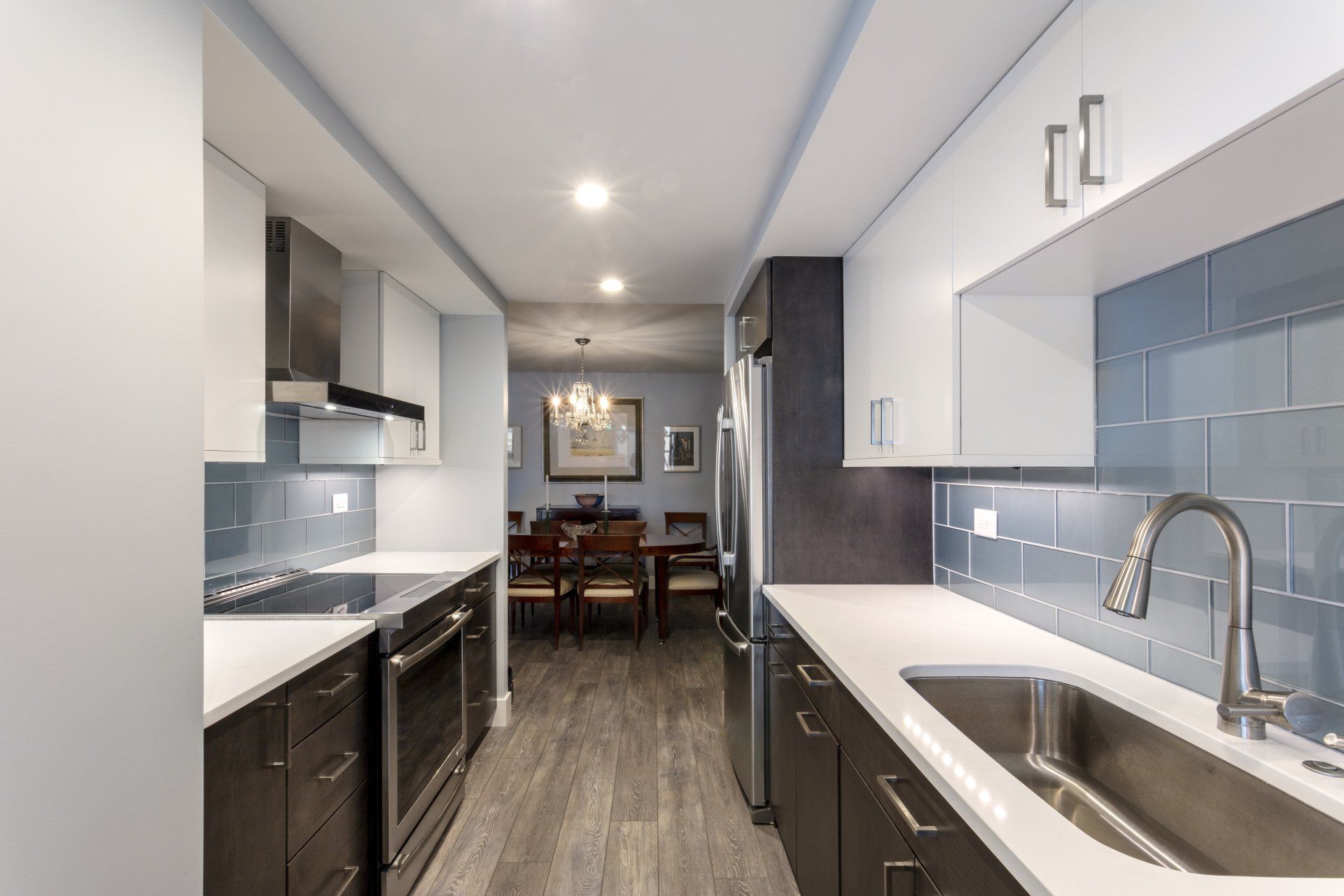 Narrow modern kitchen with white cabinets, dark counters, stainless appliances, and a dining area beyond.