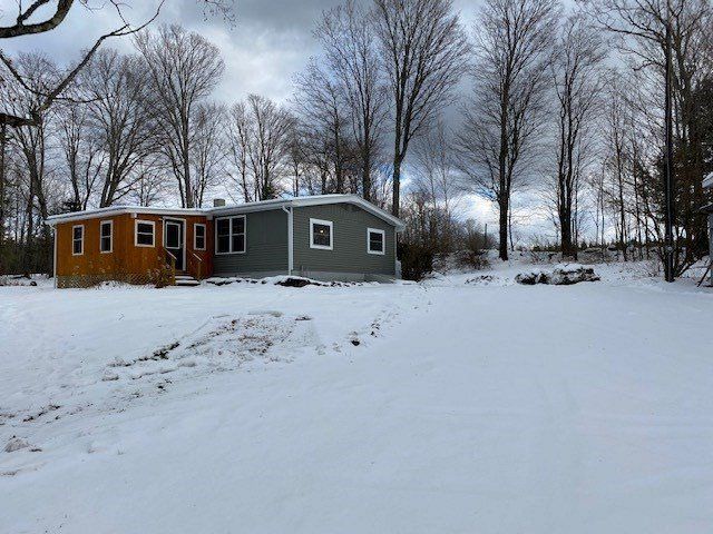 Snow-covered yard with a single-story house. The house is half wood and half gray. Bare trees in the background.