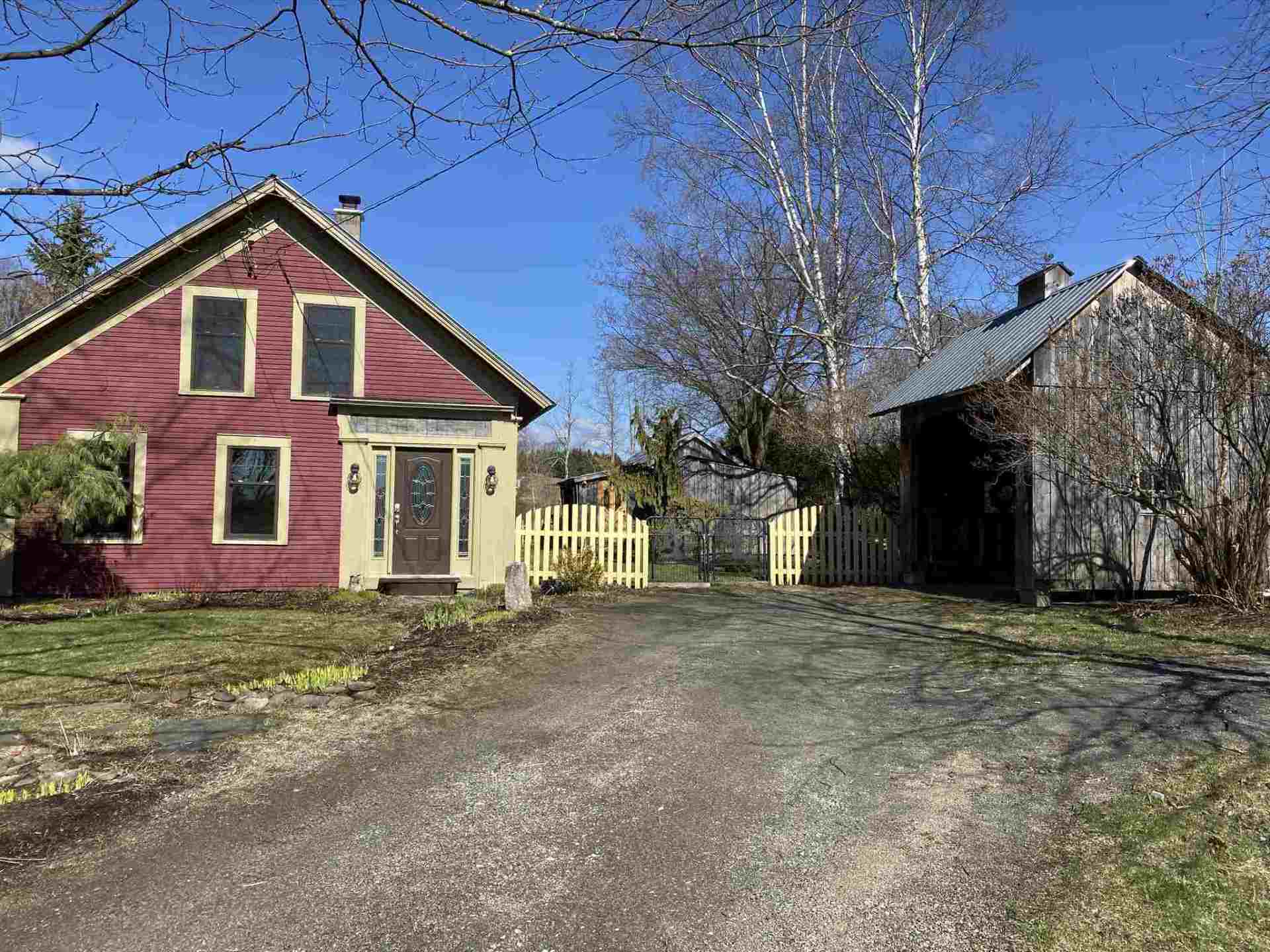 Red house with yellow trim and a gravel driveway next to a small wooden shed, under a blue sky.