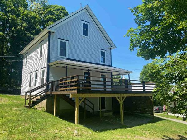 Two-story house with a wooden deck and railing. Green lawn slopes downward. Sunny day.