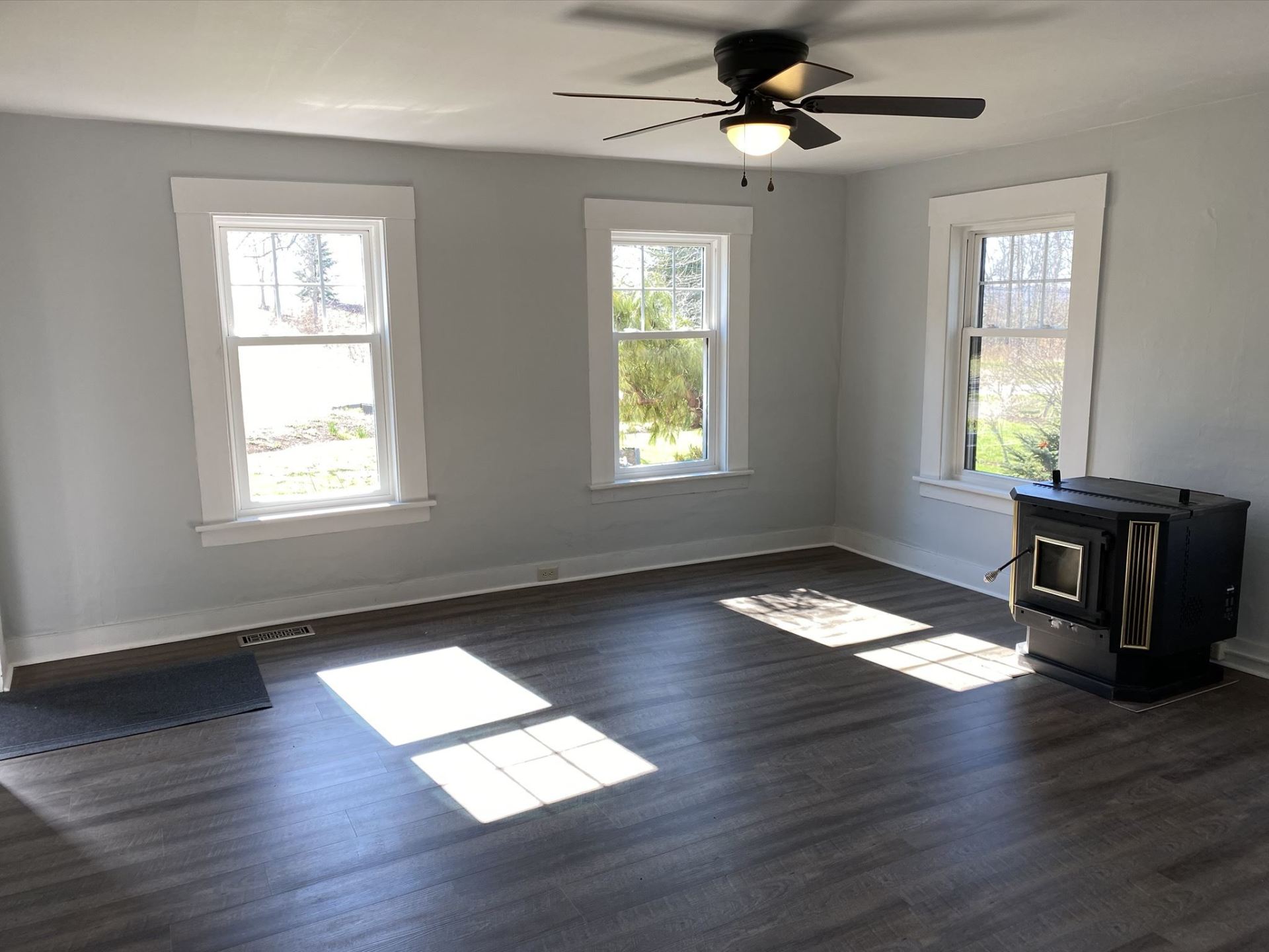 Empty living room with three windows, gray walls, and a dark wood-look floor. A black stove is in the corner.
