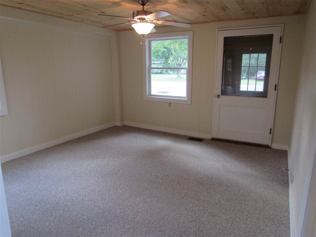 Empty room with carpet, window, and door. Ceiling fan hangs from wood-paneled ceiling.