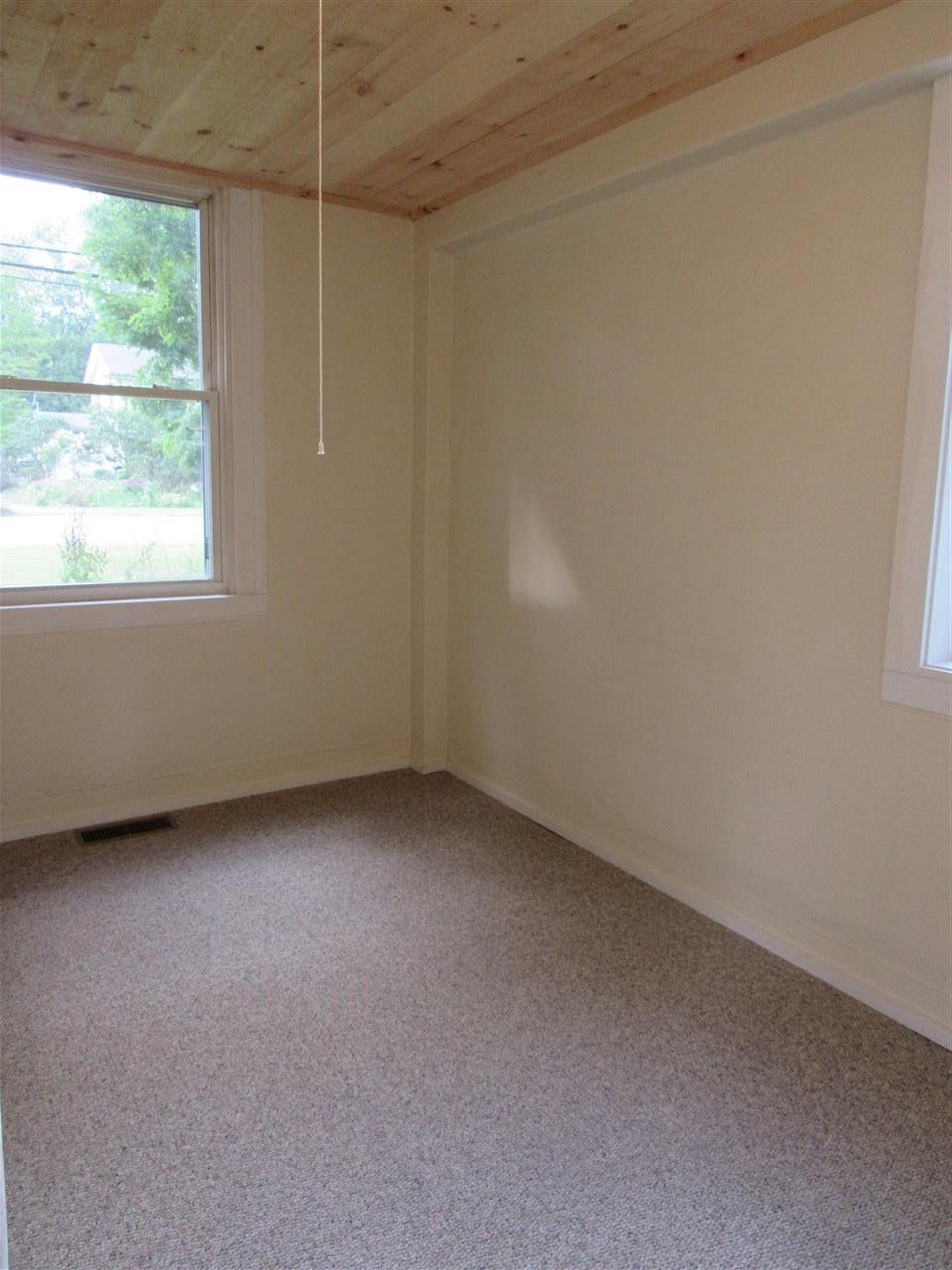 Empty room with beige walls, brown carpet, and a wooden ceiling. A window provides natural light.
