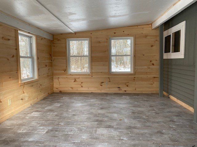 Empty room with wood-paneled walls, gray flooring, and three windows offering a view of snowy trees.