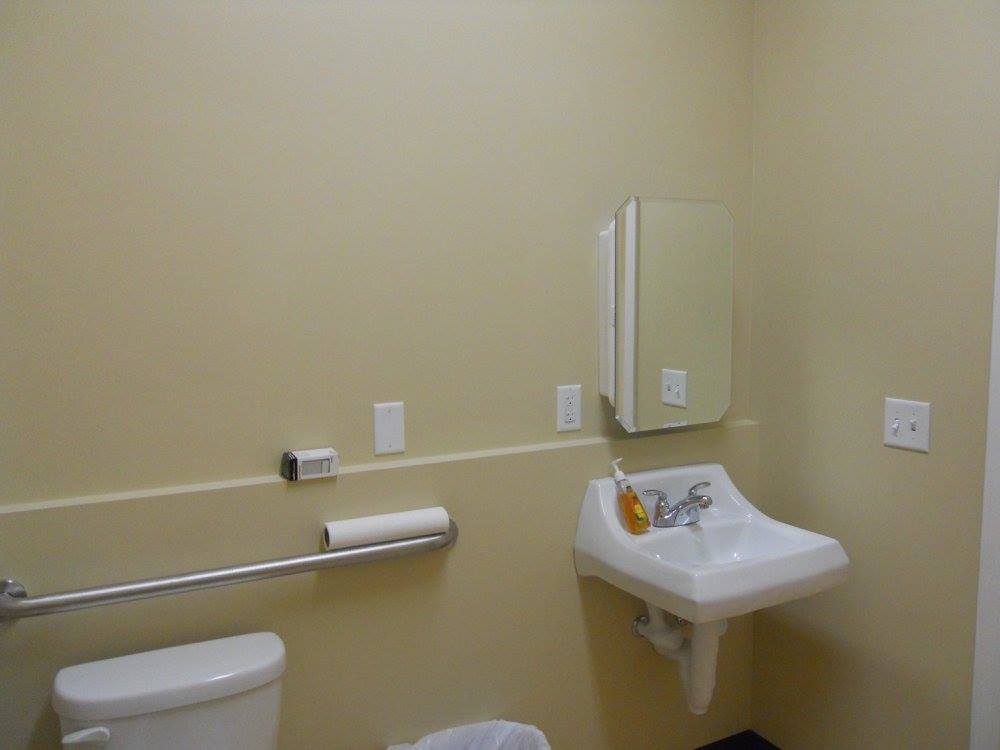 Accessible bathroom sink and toilet with grab bars, beige walls, and a wall-mounted mirror.
