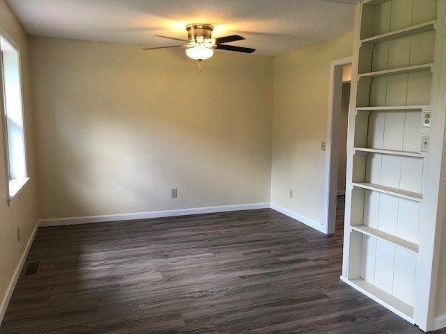 Empty living room with wood-look flooring, light-colored walls, ceiling fan, and built-in white bookshelf.