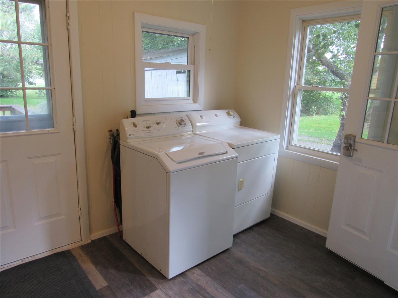White laundry machines sit under a window between two white doors, in a room with wood-look flooring.