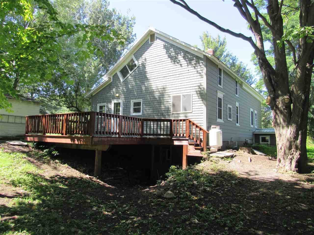 Back of gray clapboard house with wooden deck, propane tank, and overgrown yard.