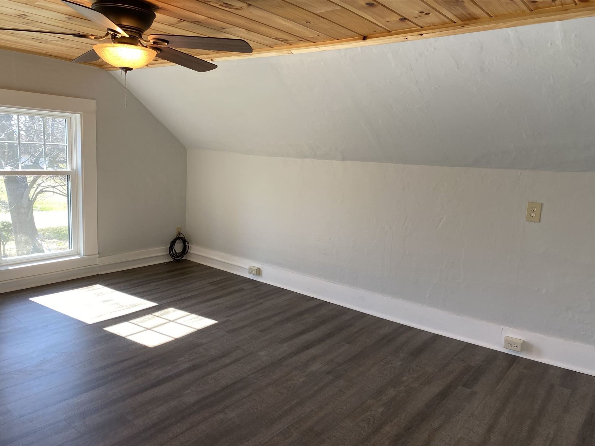 Empty room with gray walls, wood ceiling, and dark floor. Window on the left, power outlets along the base of the wall.
