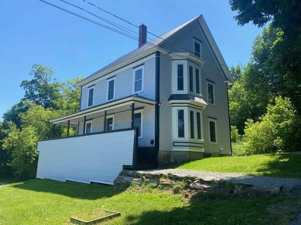 Gray house with white trim, porch, bay window, and a sloping lawn on a sunny day.