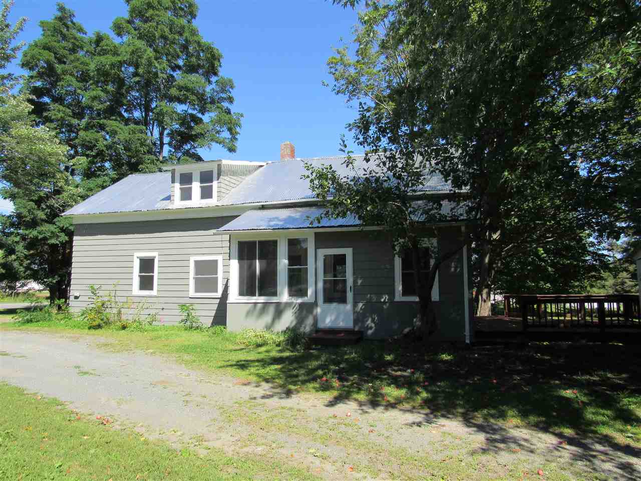 Gray, weathered house with a metal roof on a sunny day. Trees and a gravel driveway.