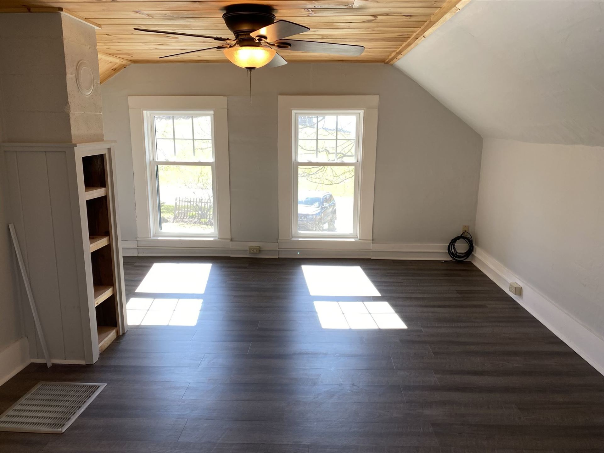 Spacious attic room with gray walls, two windows, wood floor, and wooden ceiling with a fan.