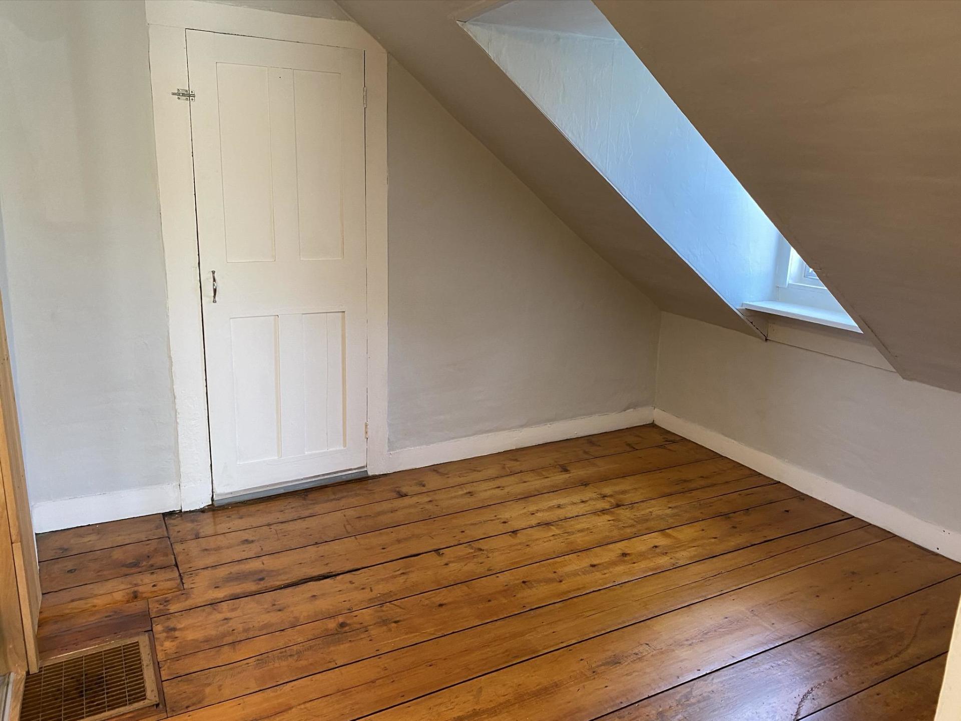 Empty room with slanted ceiling, wooden floor, white door, and small window.