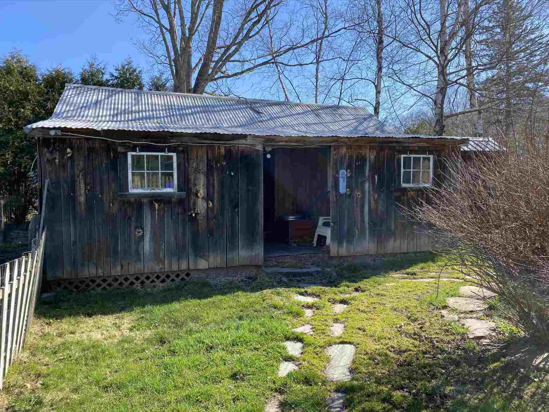 Weathered wooden shed with a corrugated metal roof and two windows. A stone path leads to an open doorway.
