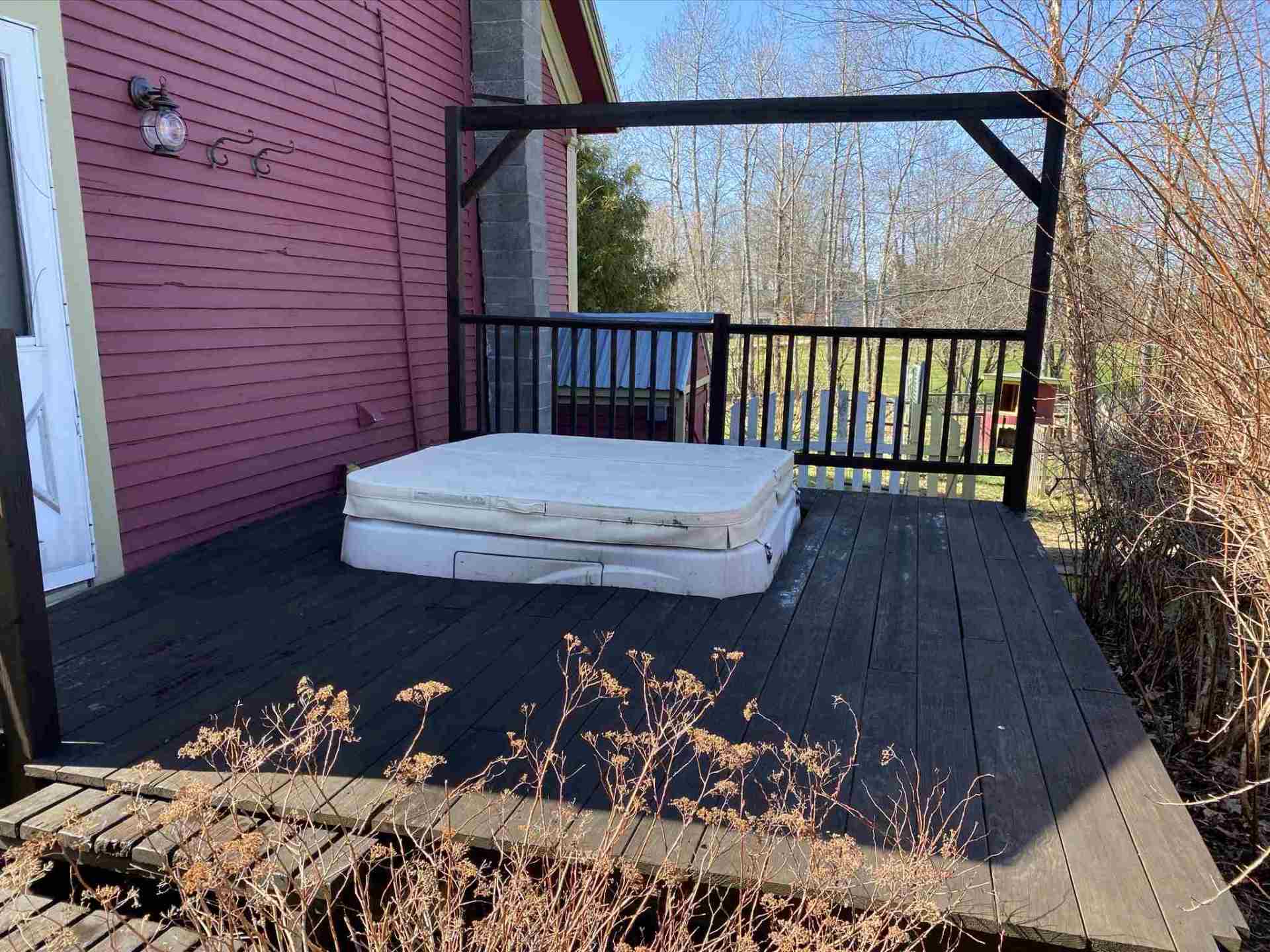Deck with a mattress on it, next to a red building and a wooded area.