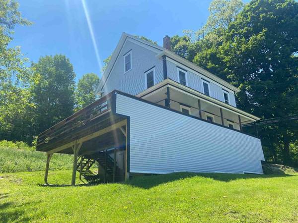 Two-story house with a large deck, surrounded by green grass and trees, under a bright blue sky.