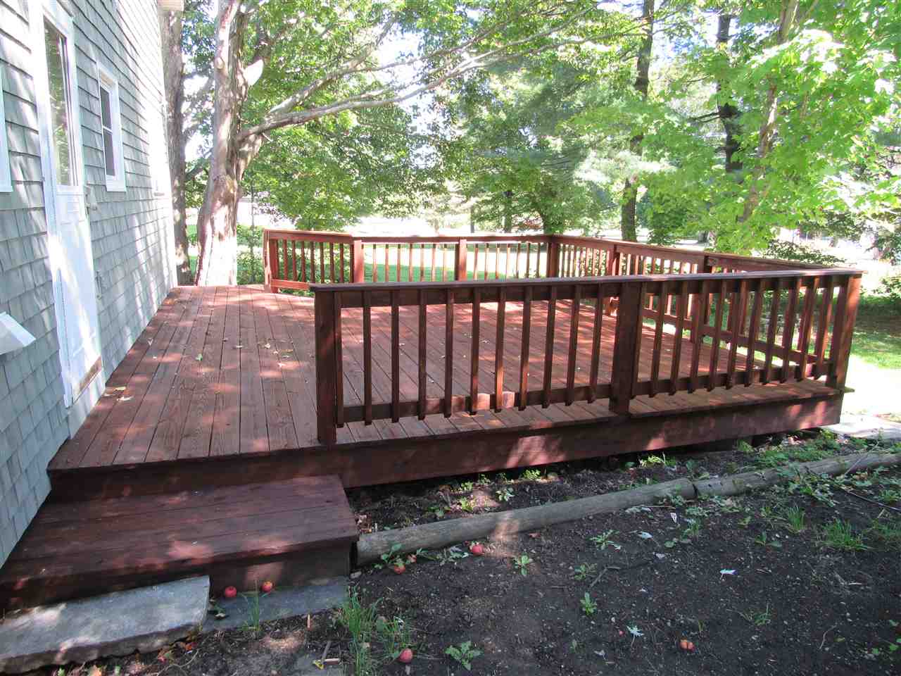 Wooden deck with brown railings attached to the side of a gray house, surrounded by grass and trees.