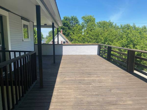 Wooden deck with dark railing and support posts, next to a light-colored house. Green trees and blue sky visible.