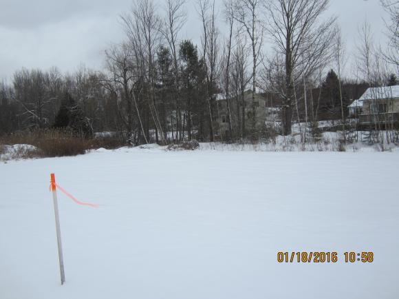 Snow-covered field with a marker, trees, and buildings in the background under a cloudy sky.