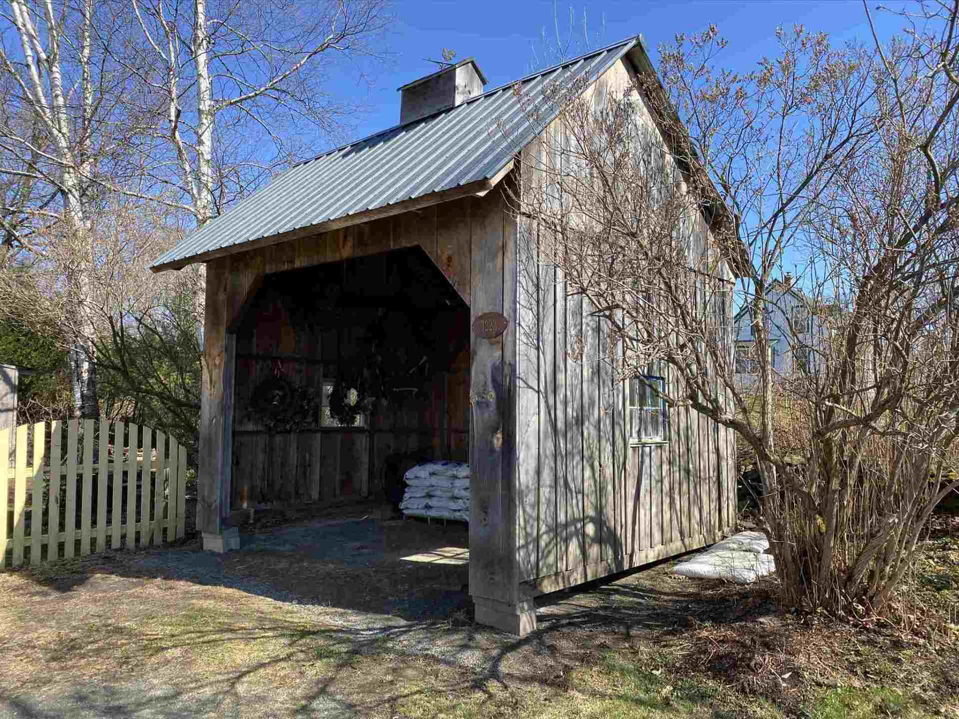 Wooden shed with a corrugated metal roof, next to a yellow picket fence, on a sunny day.