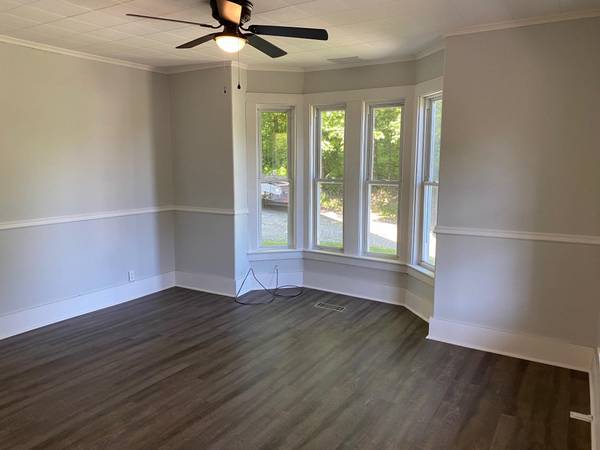 Empty room with gray walls, white trim, bay window, dark wood-look floor, and ceiling fan.