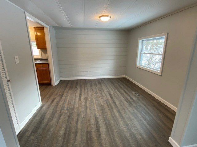 Empty room with gray walls and wood-look floor, a window, and an open doorway to a kitchen.