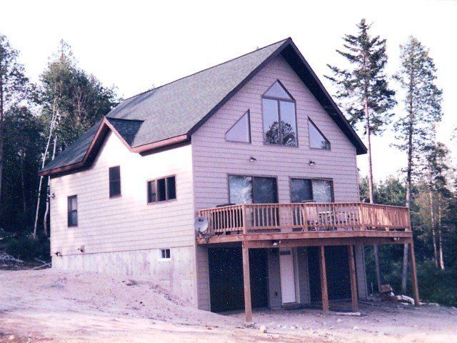 A two-story house with light-colored siding, a peaked roof, a wooden deck, and a concrete foundation in a wooded area.
