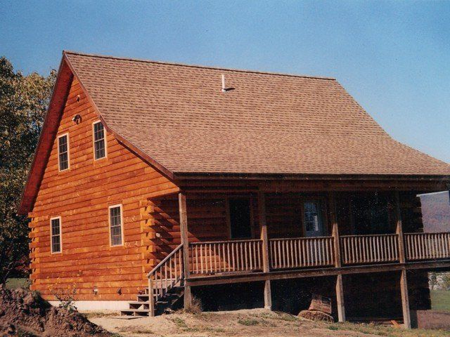 A rustic brown log cabin with a shingled roof and a covered porch stands in a grassy area under a clear blue sky.