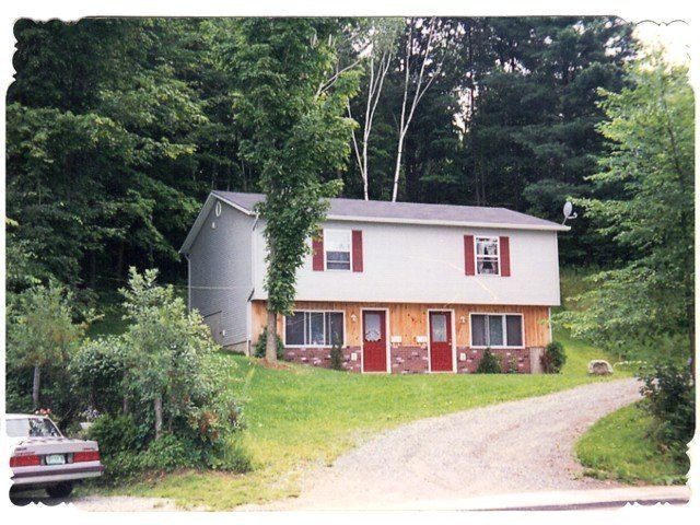 Two-story duplex with red doors and shutters, surrounded by trees on a grassy hill; a car is parked nearby.