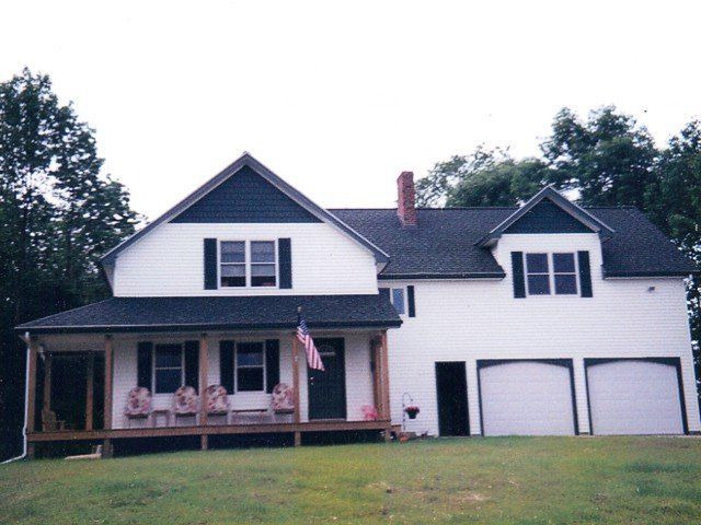A two-story white house with dark shutters, a front porch, and an attached two-car garage under a cloudy sky.
