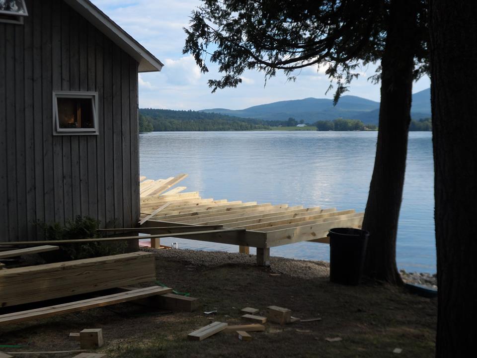 Wooden dock by a calm lake beside a gray cabin, with mountains and trees in the distance.