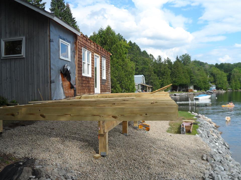 Wooden dock by a dark boathouse on a rocky lakeshore with trees and boats in the distance