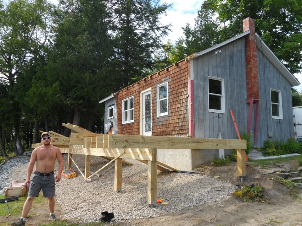 Man standing beside a partially built wooden deck on a rustic cabin in the woods