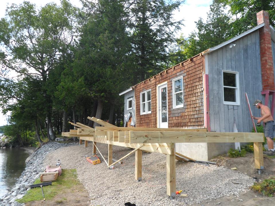House under renovation beside water, with wooden ramp and scaffolding on a gravel shoreline