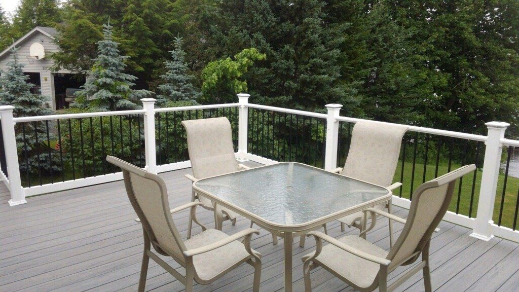 Patio with glass-top table and four chairs on a deck overlooking green trees and a yard