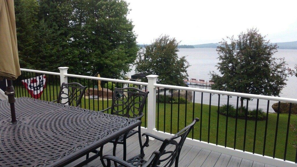 Patio table and chairs overlooking a grassy yard, trees, and a calm lake beyond a railing