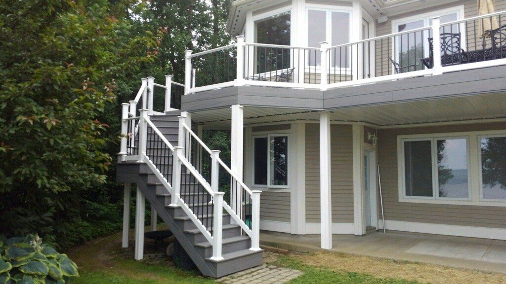 White two-story house with a wraparound porch and exterior stairs beside a wooded yard