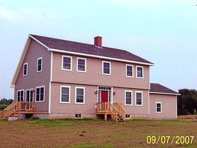 Two-story beige house with brown roof and wooden porches on a grassy lot under a clear sky