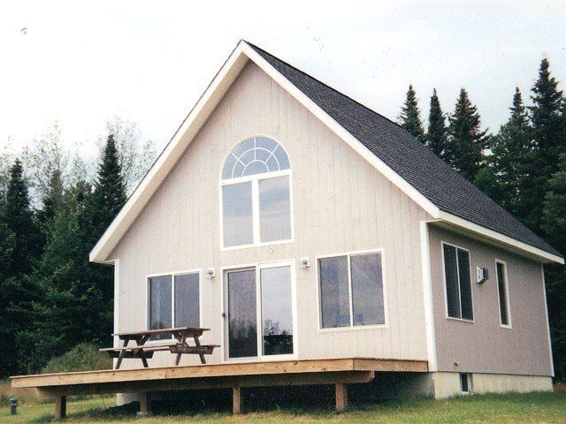 A light-colored A-frame cabin with a wooden deck and picnic table, situated in a grassy clearing surrounded by trees.