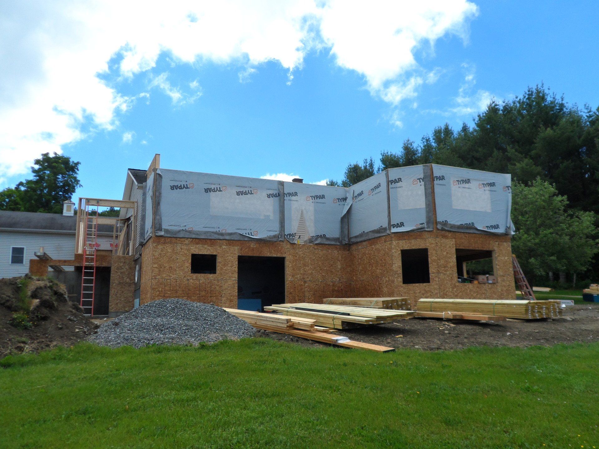 Partially constructed house with white siding, plywood lower walls, and building materials in front under a blue sky