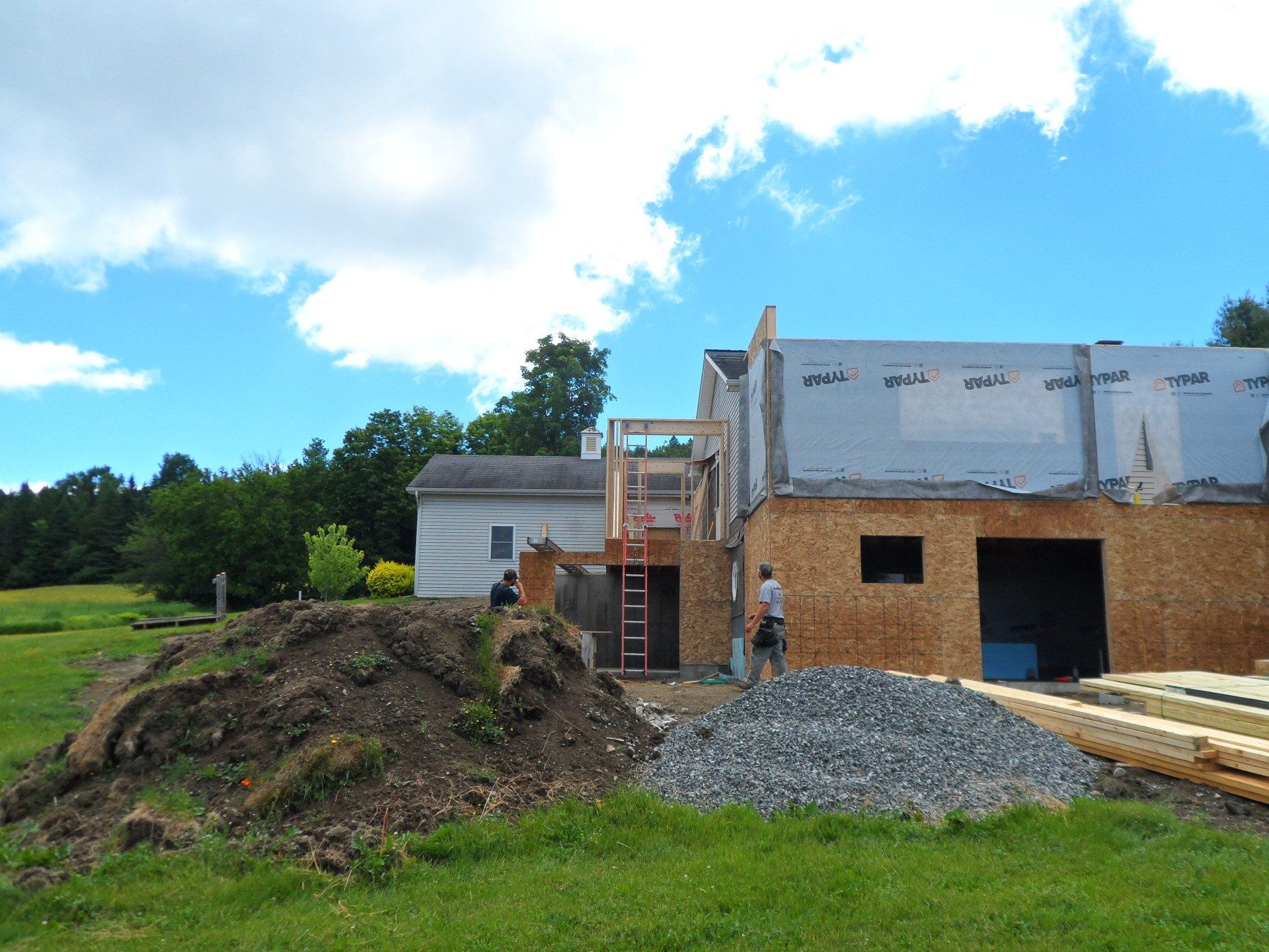 Construction site with a partially built house, gravel pile, and workers under a blue sky.