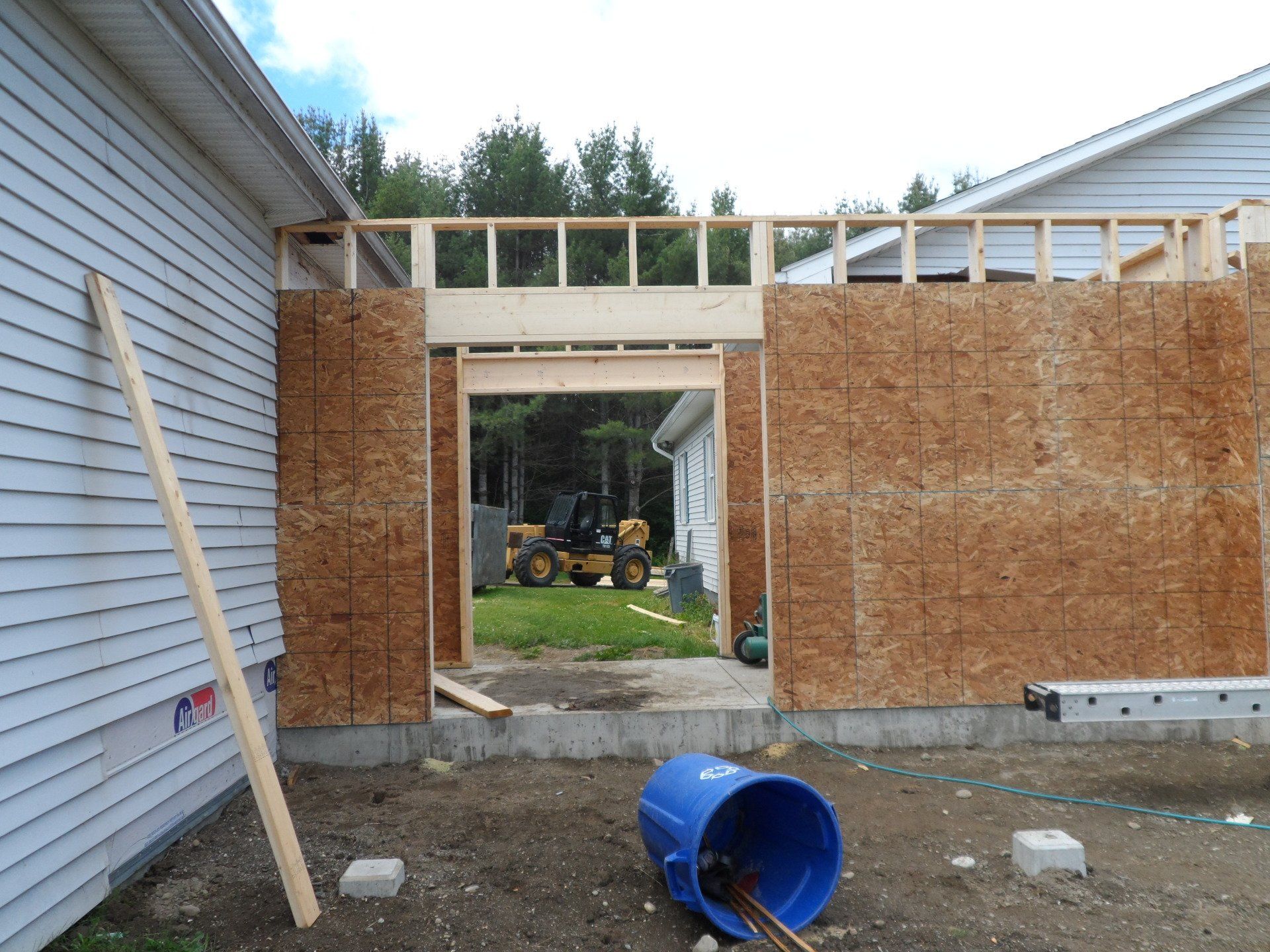 Partially built wall beside a house with exposed framing, plywood panels, and a blue bucket in the foreground