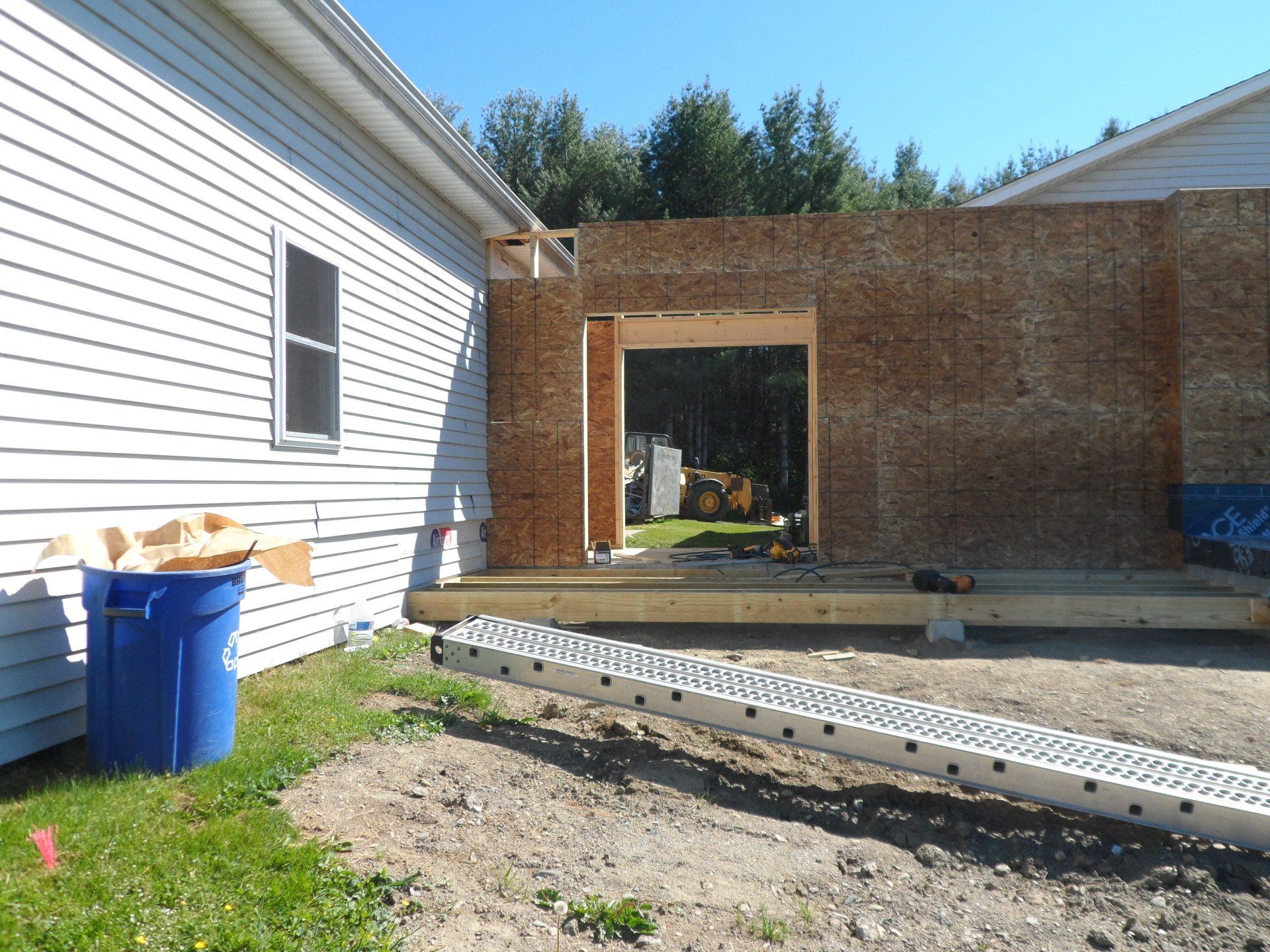 House addition under construction beside a white home, with a blue trash bin and exposed wood framing.