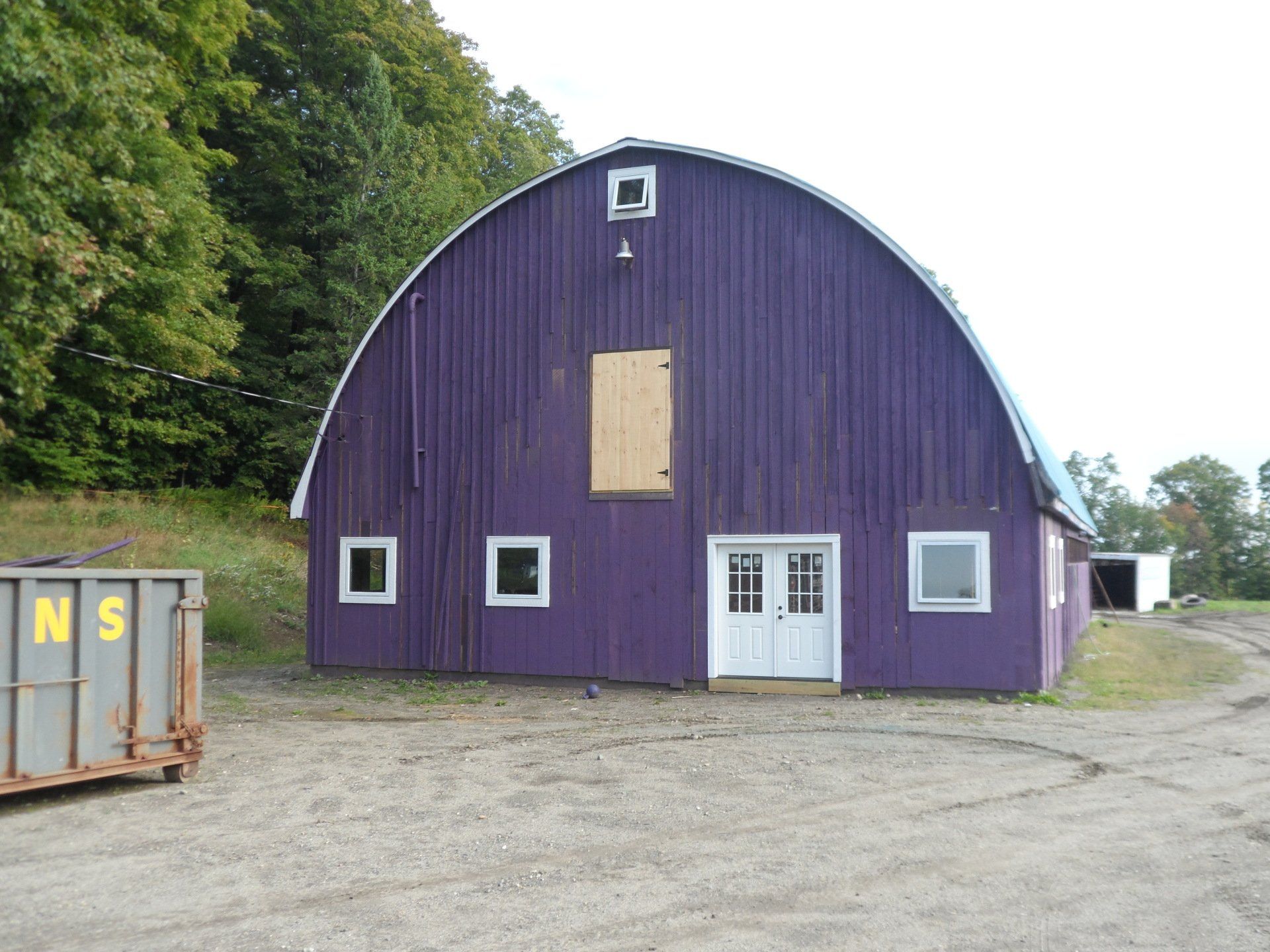 Purple arched barn with white doors and windows, set in a gravel area with trees in the background.